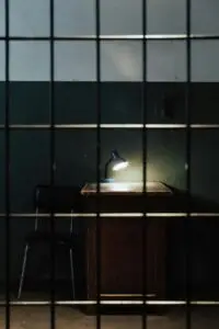 A dimly lit prison cell with a lamp on a table behind bars.